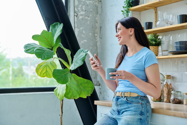 厨房里，一位美丽的女子在家中，一边喝着咖啡一边使用智能手机。这张照片捕捉了她悠闲的居家时光，展现了现代生活方式的融合。图片下载