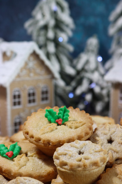 近景图片，圣诞村场景，背景是成堆的独立肉馅饼，节日的圣诞甜点，酥皮点心，甜肉馅，撒上糖粉，绿色和红色的翻糖糖霜冬青叶和浆果，姜饼屋，松树图片下载