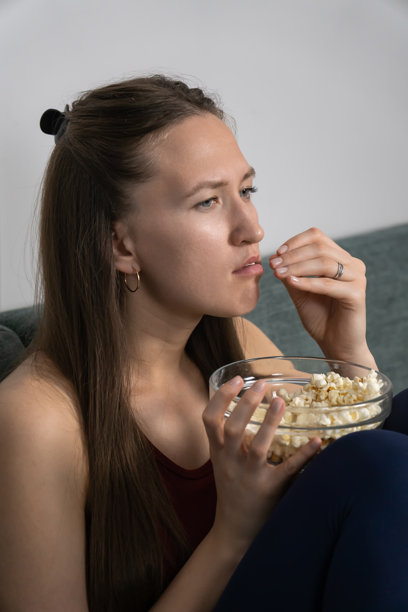 年轻女子坐在沙发上，从玻璃碗里吃着爆米花，全神贯注地观看一部引人入胜的电影，沉浸在屏幕上演绎的动作之中。竖幅照片图片下载