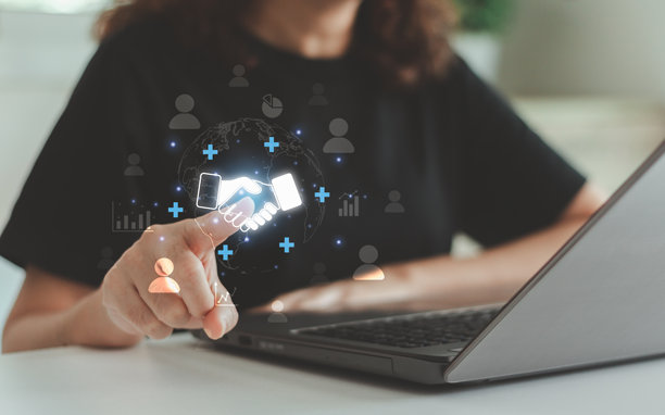 女性使用笔记本电脑，屏幕上显示数字握手全息图图标，象征在线商业合作、团队合作、虚拟协作和全球互联，体现现代科技与数字网络概念。图片下载
