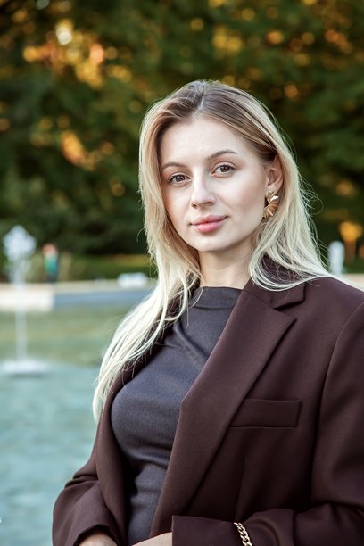 Realistic photo of a young attractive blonde businesswoman standing near a city fountain in autumn. Stylish woman in a burgundy blazer, confident and elegant, enjoying a sunny fall day outdoors.图片下载
