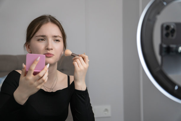 年轻的美妆博主正在用化妆刷上妆，同时用智能手机和环形灯为在线观众录制美妆教程。图片下载