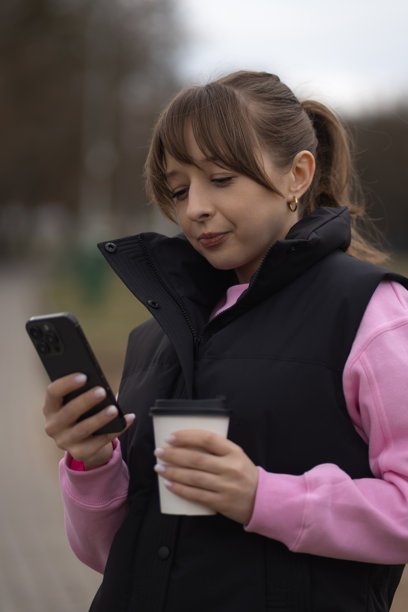 年轻女子在户外使用智能手机，手持纸杯享用咖啡，拥抱城市环境中的连接与活力。竖幅照片图片下载
