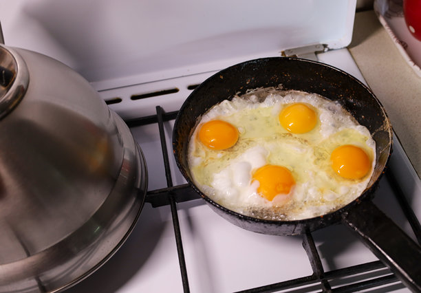 这张居家照片展示了煤气灶上的黑色平底锅里煎着四个蛋黄，旁边还有一个闪亮的烧水壶。这张照片象征着早餐准备、家庭舒适和食物，营造出一种诱人且逼真的静物画面。图片下载