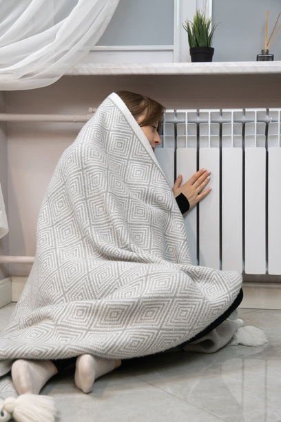 女人裹着毯子感到寒冷，触摸散热器取暖，象征着家庭环境中不断上涨的能源成本和冬季供暖问题。竖拍照片图片下载