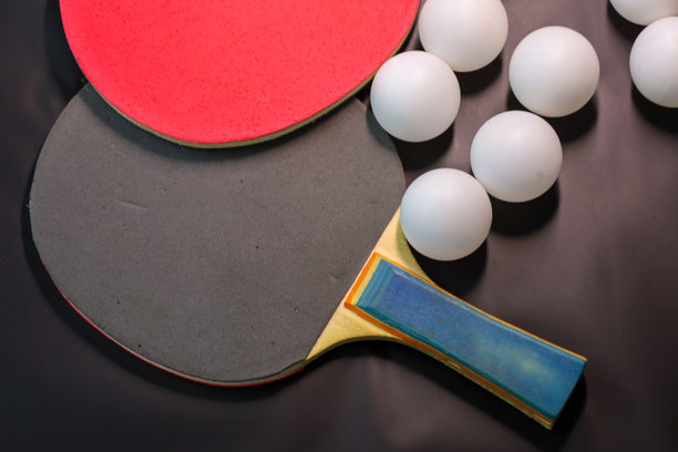 近景展示了乒乓球运动器材——两个带有红黑胶皮的球拍和几个白色的乒乓球。在深色背景下，这张图片形成了鲜明的对比，象征着比赛、竞争和健康的生活方式。图片下载