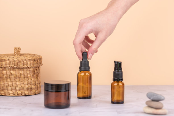 女性的手在纹理石材台面上拿起琥珀色喷雾瓶，旁边是化妆品罐和柳条篮，背景为桃色。极简风格的横向图片，留有复制空间。概念为环保的自我护理和慢美学。图片下载