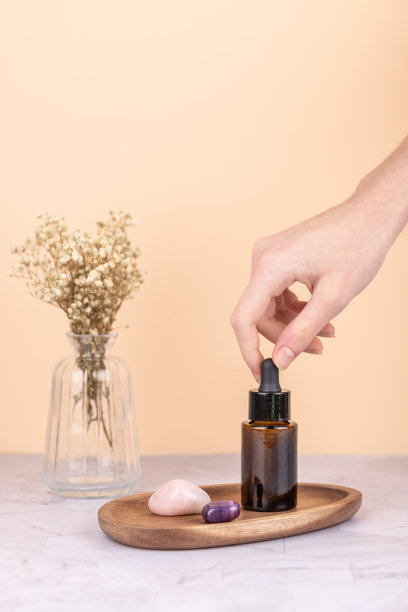 女性手持琥珀滴管瓶，置于装有干花的玻璃花瓶和装饰石头的木质托盘上。背景为桃色，表面为大理石。图片为竖幅，留有复制空间。主题为自我护理仪式和纯净美妆。图片下载