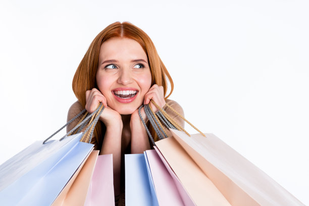 照片：一位穿着米色衬衫的有趣、兴奋的红发女士，双手托着脸颊，手里拿着购得的商品，目光望向空白的虚空，背景为纯白色。图片下载