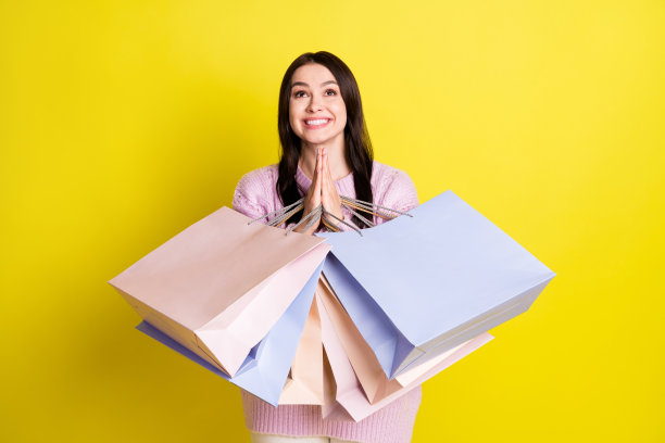 照片：一位年轻女士穿着毛衣，提着购物袋，孤立在黄色背景上，她双手合十，表情恳求。图片下载