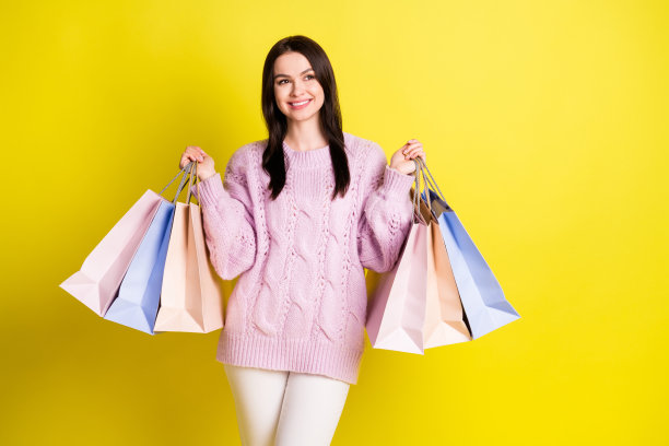 照片：一位酷炫的千禧一代女士拎着购物袋，享受购物乐趣，看向空白空间，身穿毛衣，背景为黄色。图片下载