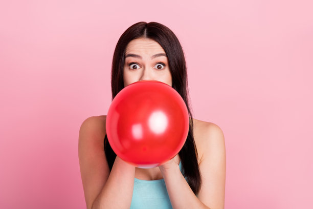 照片：一位印有千禧一代的女士，吹气球，穿着蓝色上衣，孤立在粉色背景上，表情惊讶。图片下载