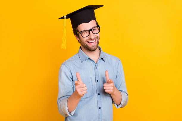 照片：一个年轻男子带着快乐的微笑，眨着眼睛，伸出手指指向你，做出选择和决定，戴着毕业帽，孤立在黄色背景上。图片下载