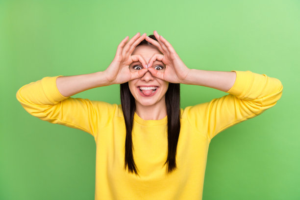 照片肖像，一位女士伸出舌头，双手拿着望远镜，孤立在鲜艳的绿色背景上。图片下载