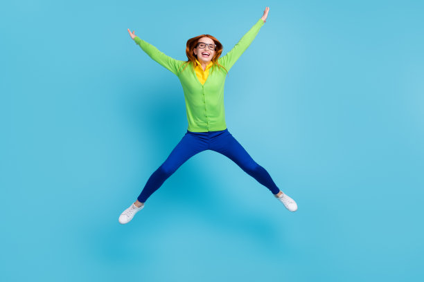 戴着眼镜、穿着开衫和裤子的红发年轻女子，在蓝色背景前跳跃，拍摄了她全身的照片，表情滑稽。图片下载