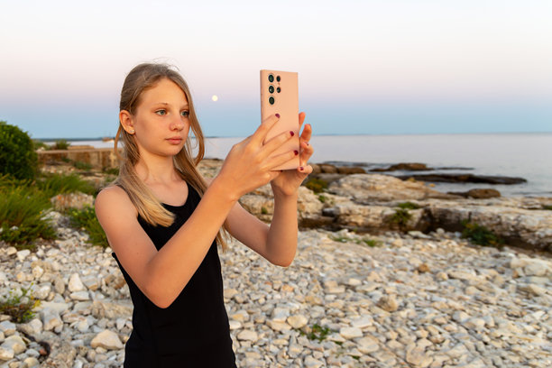 年轻女孩在岩石海滩用手机拍照。一位十几岁的少女在日落或月升时分，在海浪平静的背景下，于岩石海岸区域用智能手机捕捉照片。图片下载