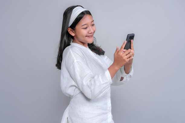 一位身穿白色衬衫、头戴发带的快乐亚洲年轻女子，正对着手机屏幕露出喜悦和兴奋的笑容，背景为纯灰色。图片下载