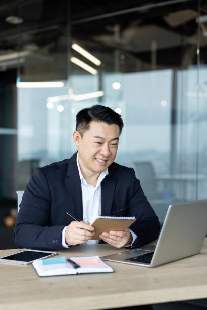 一名亚洲年轻男子在办公室使用笔记本电脑工作的垂直照片，他对着屏幕微笑，并在笔记本上做笔记图片下载