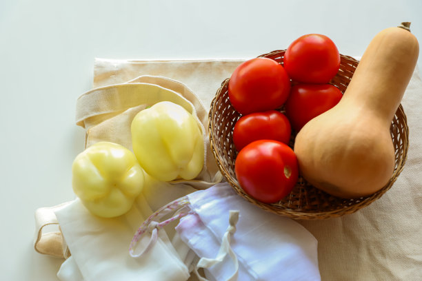 柳条篮和棉布托特包里的新鲜番茄、黄甜椒和南瓜，配以可重复使用的网袋。该照片重点展示了环保购物、健康饮食和零浪费。图片下载