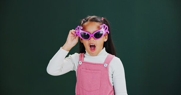 戴着太阳镜的时尚女孩在影棚摆出惊奇表情，展现时髦风格或新闻感，背景为绿色。这张肖像照片捕捉了一个孩子戴着时尚太阳镜，脸上露出惊叹或惊讶的表情，适合用于展示配饰或反应，并留有空白空间进行模拟。图片下载