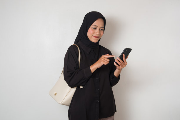 一位面带温柔微笑、身着黑色头巾的快乐亚裔穆斯林女性正在随意地使用她的智能手机。照片为影棚拍摄，背景明亮且中性。图片下载