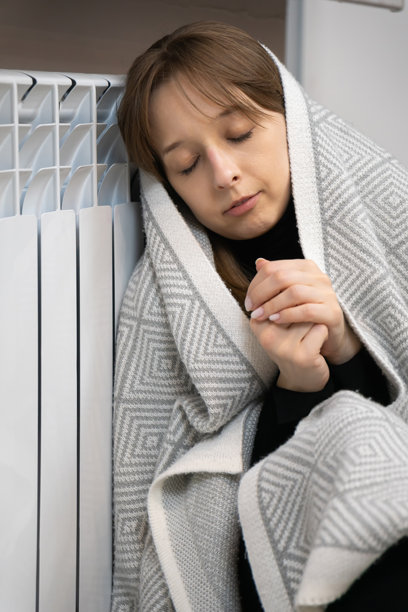 一名女子在冬天室内，裹着舒适的毯子，因寒冷的天气和高昂的能源价格感到寒冷，她触摸家里的暖气片取暖。竖屏照片图片下载