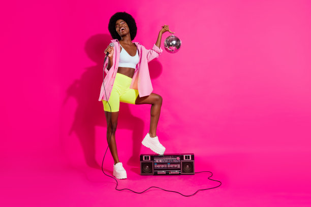 照片：可爱的、有趣的深肤色女士穿着休闲衬衫，站着，拿着音响，唱着歌，手里拿着迪斯科球，背景是孤立的粉红色。图片下载