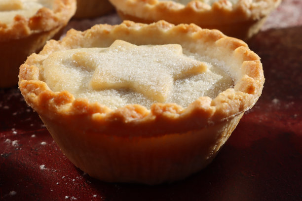 全画幅图片，展示了成排的自制肉馅派，顶部有星星状的盖子，这是节日圣诞节的甜点酥皮糕点，内有甜肉馅，撒上糖粉，背景为斑驳的红色，焦点在前景。图片下载