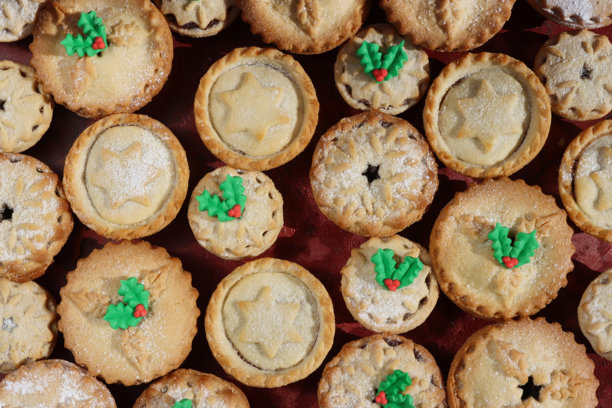 全画幅图片，展示了圣诞肉馅饼，上面装饰着糕点星星、雪花或冬青叶形状的盖子，以及绿色和红色的翻糖冬青叶和浆果。这款节日圣诞甜点采用酥皮糕点制作，内含甜肉馅，并撒上了糖粉，背景为红色。图片下载