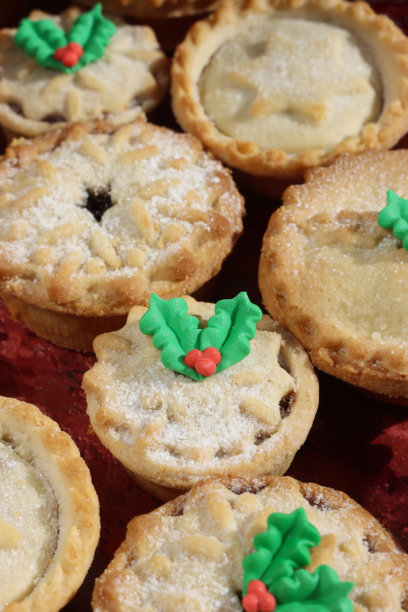 全画幅图片，展示了上面装饰有酥皮星星、雪花或冬青叶细节盖子的肉馅饼，以及绿色和红色的翻糖冬青叶和浆果，节日的圣诞甜点酥皮肉馅饼，撒上糖粉，背景为红色。图片下载