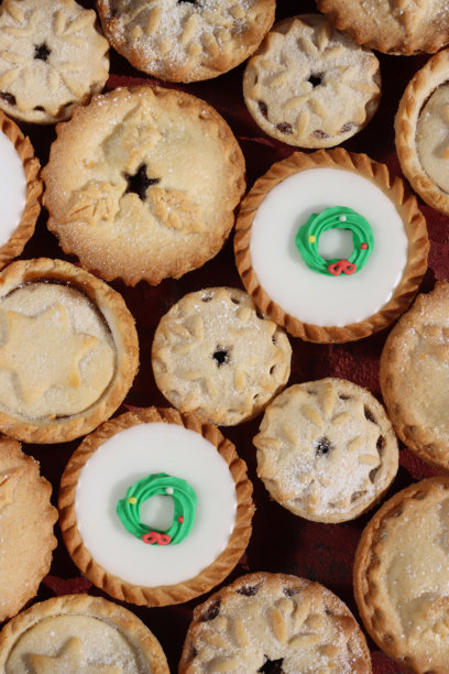 全画幅圣诞肉馅饼图片，顶部装饰有白色糖霜或糕点星星、雪花或冬青叶图案的盖子，节日圣诞甜点酥皮点心，内含甜肉馅，撒有糖粉或饰有绿色和红色糖霜圣诞花环，俯视拍摄图片下载