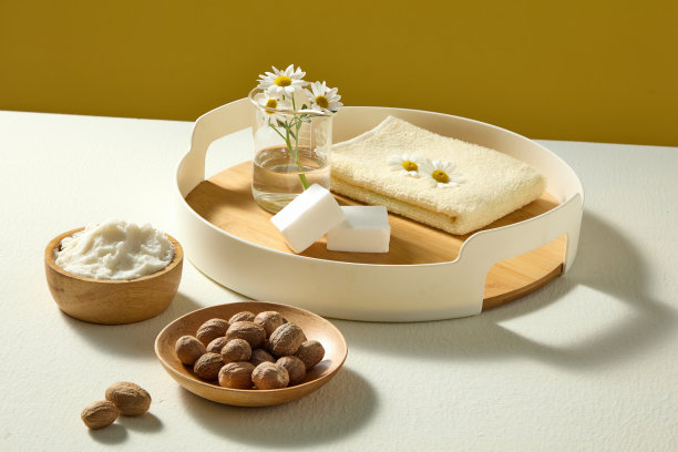 俯视木盘中乳木果油块，木盘上放有坚果和洋甘菊，背景为白色表面和黄色，适合化妆品产品设计。图片下载