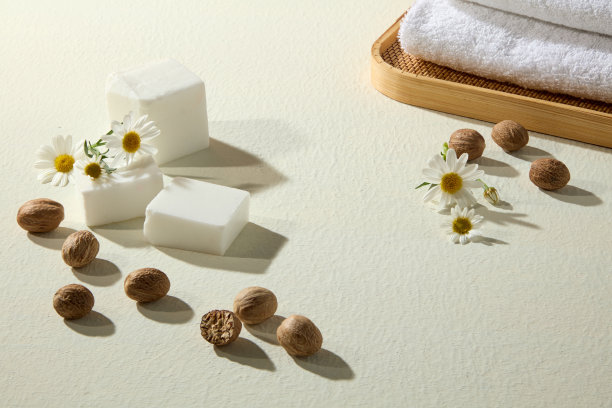 白色背景上摆放着乳木果油方块、散落的坚果、柔软的毛巾和洋甘菊，极简风格，适合化妆品品牌宣传。图片下载