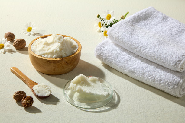 木碗盛着奶油状的乳木果油，配有坚果、洋甘菊、玻璃碗和柔软折叠的毛巾，在白色表面拍摄，用于自然美容视觉效果。图片下载