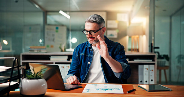 一位成熟快乐的男士在办公室的笔记本电脑前，屏幕上有海浪和夜景，可能是在进行视频通话或在线会议。这位男士可能是金融顾问，正在深夜处理统计数据、文件，或是通过网络研讨会打招呼。图片下载