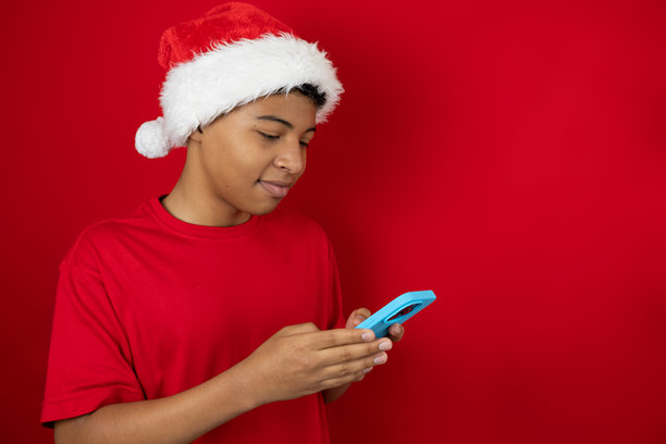 戴着圣诞帽、穿着红衬衫的快乐拉丁裔青少年，在有复制空间的红色背景上使用手机，圣诞节的年轻化风格图片图片下载