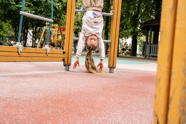 一个小女孩长发倒挂在游乐场的杆子上，脸上洋溢着顽皮的笑容。这张图片捕捉了户外环境中童年的快乐和无忧无虑的玩耍时光。图片下载