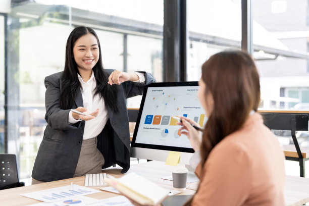 一位年轻的亚洲女商人微笑着进行商务演示，用手指着电脑屏幕上的数据图表，并与同事在现代办公室里协作。图片下载
