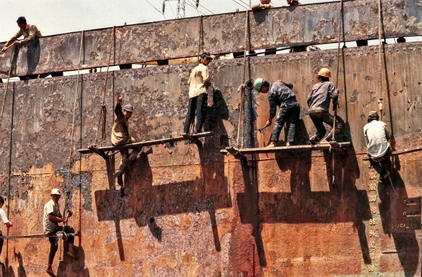 越南船厂工人在锈迹斑斑的旧船旁，在绳索悬挂的摇晃木板上高空作业拆解船只的历史彩色照片，拍摄于1992年10月20日，越南西贡。图片下载