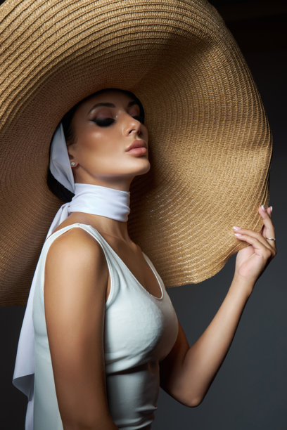 妆容精致的模特戴着宽檐草帽和时尚围巾，在光线昏暗的影棚环境中散发出优雅和精致的气息，照片捕捉了她的优雅与沉着。图片下载