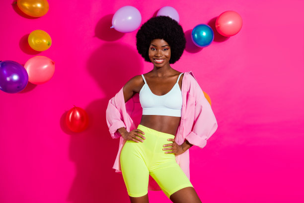 照片：一位可爱的、肤色较深的女士，身穿休闲衬衫，一手拿着漂浮的彩色气球，一手叉腰，面带微笑，背景是粉红色的。图片下载