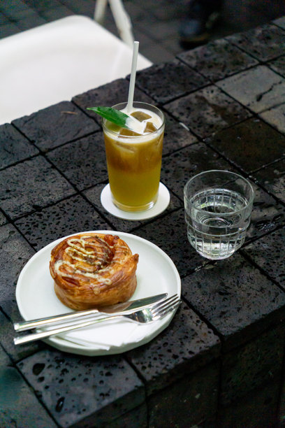 这张照片展示了一个放在白色盘子里的火腿奶酪卷，配有餐具，一杯饰有薄荷的冰镇芒果冷萃咖啡，以及放在深色湿瓷砖桌上的水杯。图片下载