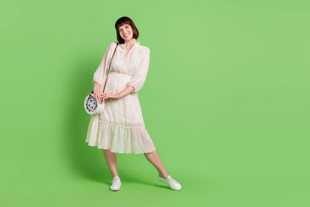 身穿波点连衣裙的微笑女子，手拿手包，全身照，在柔和的绿色背景前，全长身体尺寸照片图片下载