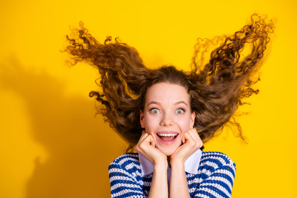 穿着条纹毛衣的年轻女子，一头蓬乱的头发，在明亮的黄色背景下兴奋地微笑，适合时尚生活方式的图库照片图片下载