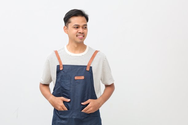 一位穿着蓝色围裙的快乐亚洲咖啡师看向镜头。咖啡店老板身穿咖啡馆制服，面带微笑，侧身站立，背景为白色。图片下载