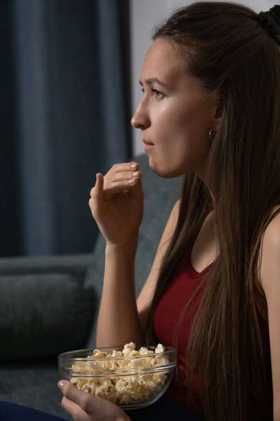 年轻女子舒适地躺在沙发上，享受着爆米花电影之夜，在宁静的夜晚沉浸于家庭影院娱乐的兴奋之中。竖屏照片图片下载