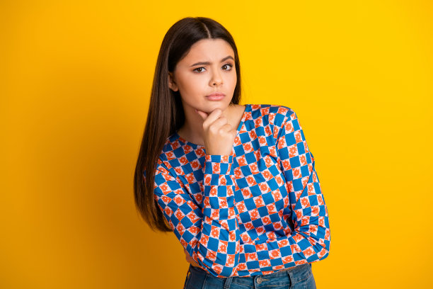 一位身穿蓝橙格纹衬衫的年轻女子，在明亮的黄色背景前若有所思地摆出造型，这是一张时尚生活类图库照片。图片下载