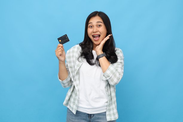 照片：一位印有年轻亚洲女性，一只手托着脸颊，手持借记卡或信用卡，面露惊讶之色，张大嘴巴开心地看着镜头，背景为蓝色。图片下载