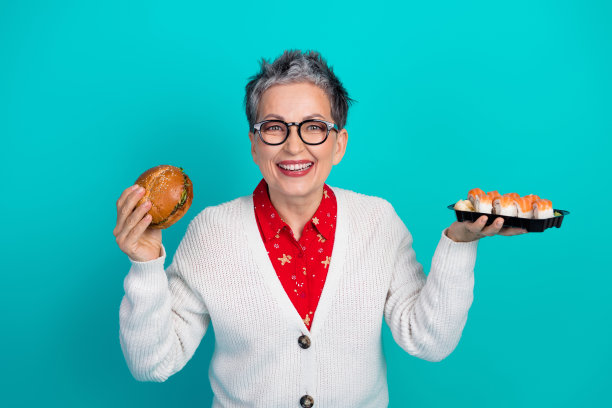 穿着开襟羊毛衫的成熟的女士面带微笑，手持汉堡和寿司托盘，背景为蓝绿色，拍摄圣诞节主题的节日照片图片下载