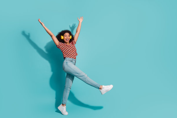身穿休闲印花衬衫、牛仔裤和白色运动鞋的年轻女子戴着黄色耳机，在蓝色背景前欣喜若狂地跳跃，生活方式照片图片下载